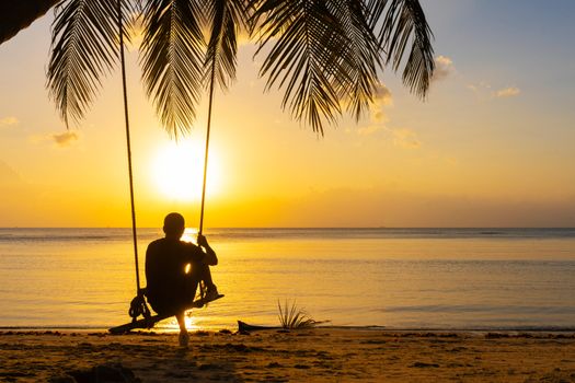 The guy enjoys the sunset riding on a swing on the ptropical beach. Silhouettes of a guy on a swing hanging on a palm tree, watching the sunset in the water