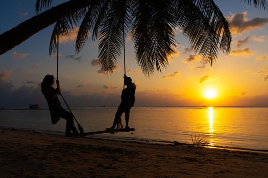 Silhouetted couple in love walks on the beach during sunset. Riding on a swing tied to a palm tree and watching the sun go down into the ocean.