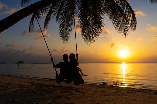 Silhouetted couple in love walks on the beach during sunset. Riding on a swing tied to a palm tree and watching the sun go down into the ocean.