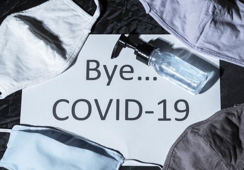 Light shines on the paper labeled Bye COVID 19 with protective face mask and gel alcohol on dark background. Concept of say goodbye for Corona virus pandemic.