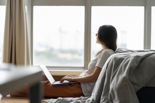 Beautiful young brunette girl working on a laptop, sitting on the floor near the bed by the panoramic window. Stylish modern interior. A cozy workplace. Shopping on the Internet