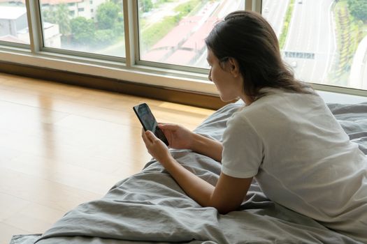 A beautiful young brunette girl is texting on the phone while lying on a bed by a panoramic window with a beautiful view from a high floor