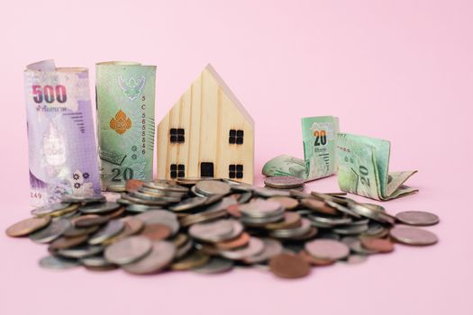 Wooden house model with Thai currency banknote and money coins on pink background for business, finance and property investment concept
