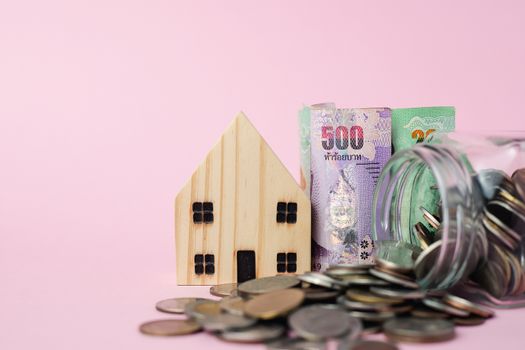 Wooden house model with Thai currency banknote and money coins in the glass jar on pink background for business, finance and property investment concept