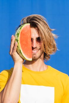 Attractive young man with long blond hair with half a watermelon covering his face on a plain blue background. Summer, sun, heat, fruit, vacation concept.