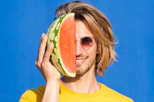 Attractive young man with long blond hair with half a watermelon covering his face on a plain blue background. Summer, sun, heat, fruit, vacation concept.