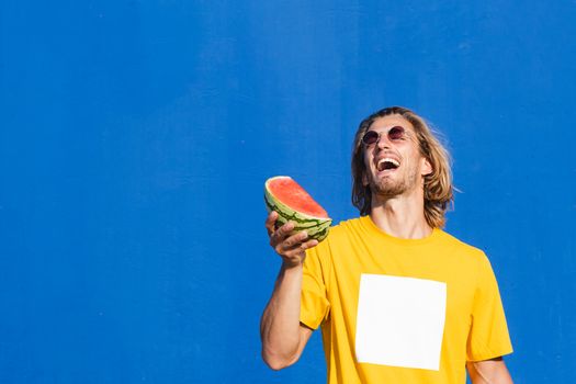 Attractive young man with long blond hair holding half a watermelon in his hand smiling and enjoying himself on a plain blue background. Summer, sun, heat, fruit, vacation concept.