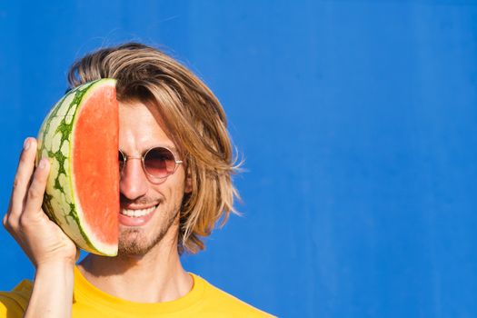 Attractive young man with long blond hair with half a watermelon covering his face on a plain blue background. Summer, sun, heat, fruit, vacation concept.