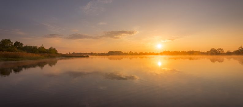 Scenic view of beautiful sunrise or dawn above the pond at spring or early summer morning with cloudy sky background and fog over water. Landscape. Water reflection.