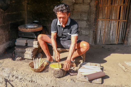 Old man is cutting the bamboo in his workshop to make bamboo straws, Bali, Indonesia. Front view to a worker who makes bamboo straws.