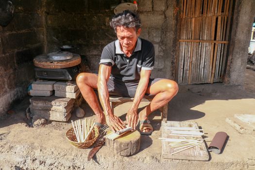 Old man working with bamboo in his workshop to make bamboo straws, Bali, Indonesia. Ecological way of producing recyclable products. Front view to a worker who makes bamboo straws.
