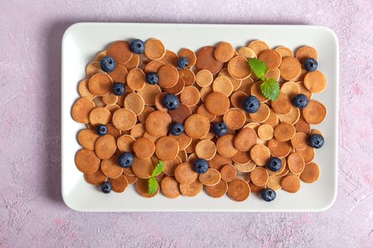 Trendy homemade breakfast, pancake cereal, mini pancakes on light plate on pink concrete background with blueberry, leaves of lemon balm and peppermint