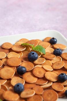 Trendy homemade breakfast, pancake cereal, mini pancakes on light plate on pink concrete background with blueberry, leaves of lemon balm and peppermint. Space for text