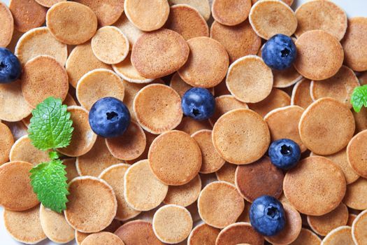 Trendy homemade breakfast, pancake cereal, mini pancakes on light plate on pink concrete background with blueberry, leaves of lemon balm and peppermint