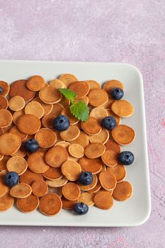 Trendy homemade breakfast, pancake cereal, mini pancakes on light plate on pink concrete background with blueberry, leaves of lemon balm and peppermint. Space for text