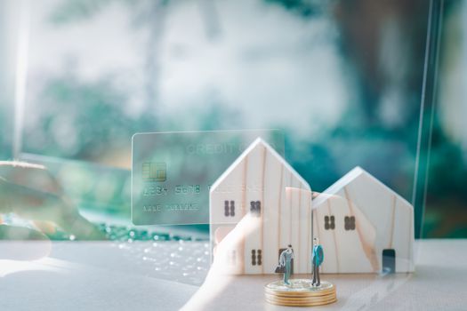 Business and financial concept. Double exposure hand holding credit card and wooden home with small people model stand on a stack of golden coins.