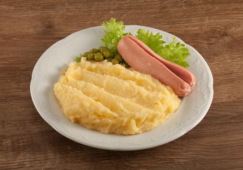 White plate with mashed potatoes, boiled small sausage, green peas and fresh green lettuce on the wooden table