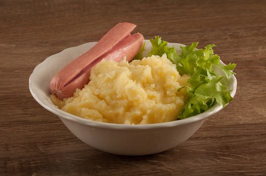 White bowl with mashed potatoes, boiled small sausage and fresh green lettuce on the wooden table