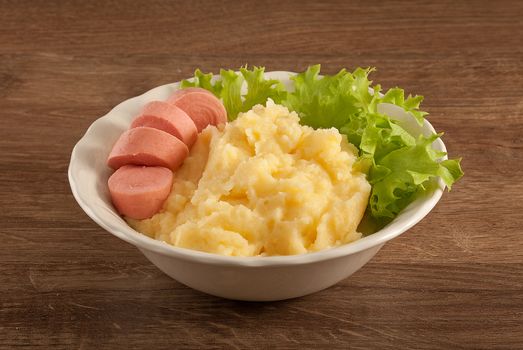 White bowl with mashed potatoes, boiled small sausage and fresh green lettuce on the wooden table