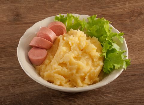 White bowl with mashed potatoes, boiled small sausage and fresh green lettuce on the wooden table