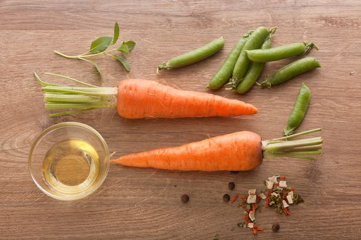 Top view of two whole carrots, fresh green pea pods, oil and dry herbs on the wooden board