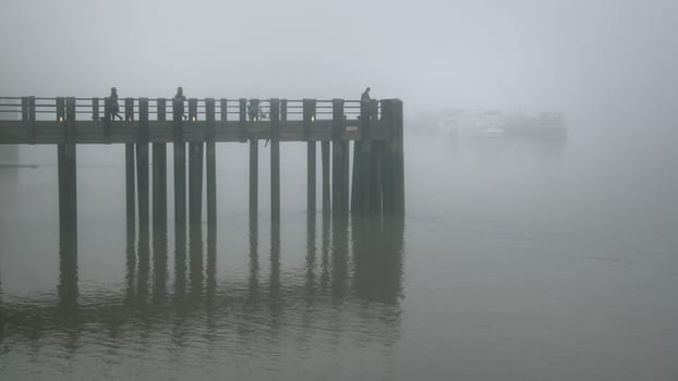 Silhouettes of people and bicycle on wooden pier on river Thames in London during dull gray foggy evening.