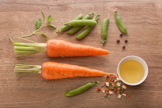 Top view of two whole carrots, fresh green pea pods, oil and dry herbs on the wooden board