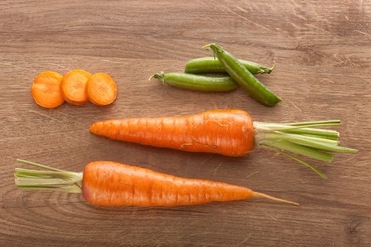 Top view of two whole carrots, some carrot's slices and fresh green pea pods on the wooden table