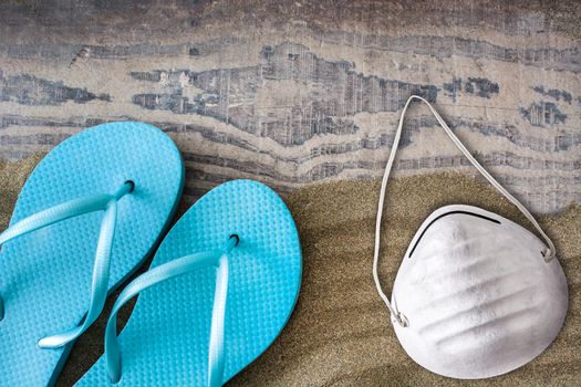 Summer shoes and protective face mask on sand background