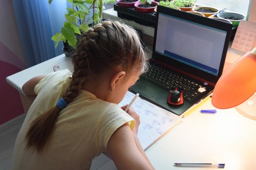 Girl solves math examples in notebook in front of laptop