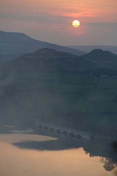 Calm Ladybower reservoir water beneath an arched bridge reflects the orange sunset from above the tall hills.