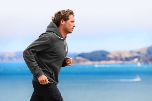 Athlete man running in sweatshirt hoodie in autumn fall by the water. Male runner training outdoors jogging in nature.