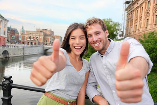 Thumbs up couple happy in Stockholm, Sweden. Excited people giving thumbs up gesture looking at camera. Multiracial young couple walking outside in Stockholm. Scandinavian man, Asian woman.