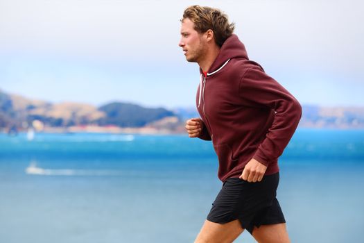 Runner athlete man running in sweatshirt hoodie in autumn fall by the water. Male runner training outdoors jogging in nature.