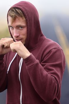Fitness boxer boxing outside on road in hoodie. Man punching and hitting looking at camera. Fit fitness boxer sweating in sweatshirt outside. Caucasian male fitness model in his 20s.