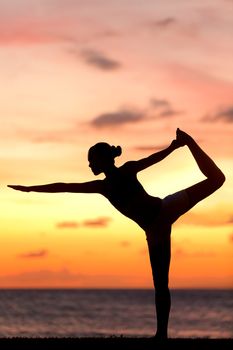 Yoga woman in serene sunset at beach doing king dancer pose. Meditation and balance exercise at sunrise or sunset with female yoga instructor exercising outside in nature.