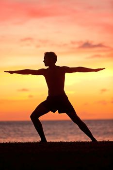 Yoga man training and meditating in warrior pose outside by beach at sunrise or sunset. Male yoga instructor working out training in serene ocean landscape. Silhouette of man model against sun.