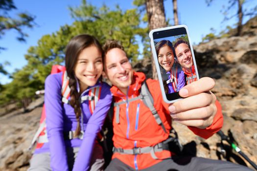 Selfie couple using smart phone hiking in nature with smartphone. Happy couple taking self-portrait photo picture using app. Man and woman having fun together. Focus on screen.