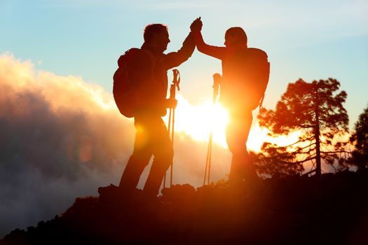 Hiking people reaching summit top giving high five at mountain top at sunset. Happy hiker couple silhouette. Success, achievement and accomplishment people