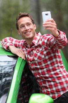 Driver by car taking selfie photo with smartphone after driving in new green car. Happy man taking picture with smartphone camera during travel road trip. Young Caucasian male model in his 20s.