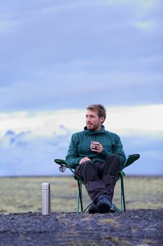 Camping man sitting in folding chair drinking coffee from thermos bottle flask at dusk in nature on Iceland. Camper relaxing thinking pensive taking break on road trip in beautiful Icelandic nature.