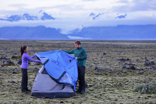 Tent - people pitching tent on Iceland at dusk. Couple setting up camp for night after hiking in the wild Icelandic nature landscape. Multicultural Asian woman and Caucasian man healthy lifestyle.