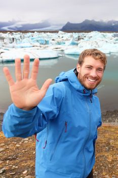 Tourist waving hand by Jokulsarlon on Iceland on travel. Portrait of happy man saying hello smiling looking at camera front of the glacial lake / glacier lagoon.
