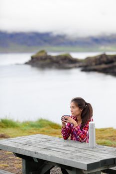 Camping woman sitting at table drinking coffee from thermos bottle flask by lake on Iceland. Camper girl relaxing thinking pensive taking break on road trip in beautiful Icelandic nature.