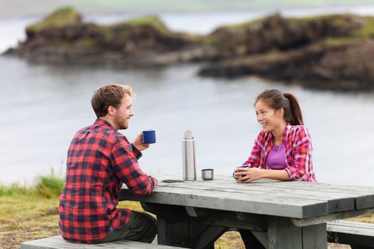 Camping couple sitting at table drinking coffee from thermos bottle flask by lake on Iceland. Campers woman and man relaxing taking break on road trip in beautiful Icelandic nature.