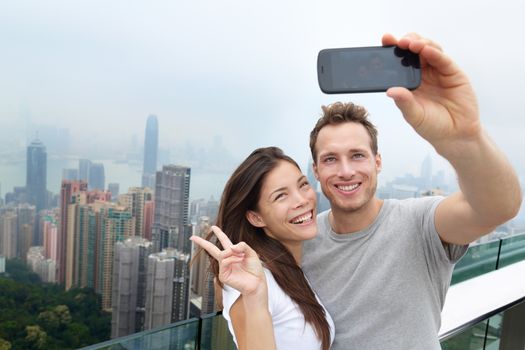 Hong Kong Victoria Peak tourists couple taking selfie photo picture with smartphone enjoying view over Hong Kong and Victoria Harbour. Young happy multiethnic couple traveling in Asia.