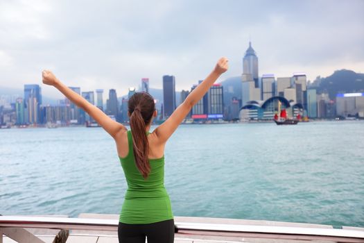 Happy woman success cheering by Hong Kong skyline with arms raised up outstretched. Successful winner celebrating cheerful on Tsim Sha Tsui Promenade and Avenue of Stars in Victoria Harbour, Kowloon.