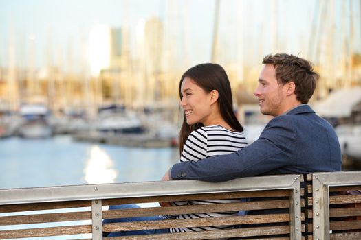 Romantic couple sitting on bench in old harbour, Port Vell, Barcelona, Catalonia, Spain. Happy woman and man embracing enjoying life and romance outside. Multiracial Caucasian Asian couple in love.