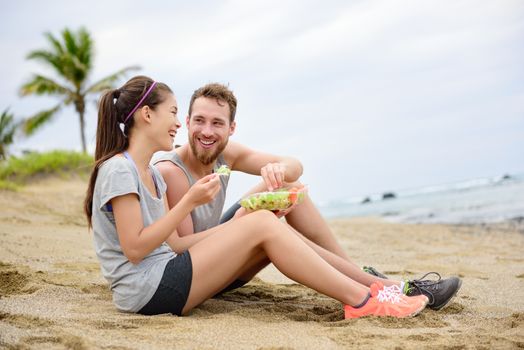 Salad - healthy fitness woman and man couple laughing eating food lunch sitting on beach after workout. Mixed race Asian Caucasian female model and male models in sportswear.