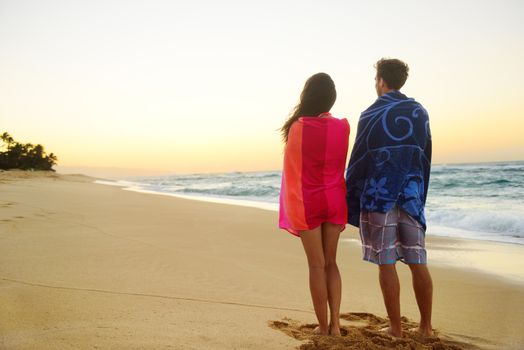 Couple standing in beach sand, towels over shoulders enjoying ocean sunset during travel holidays getaway vacation. Mixed race couple, asian woman, caucasian man.